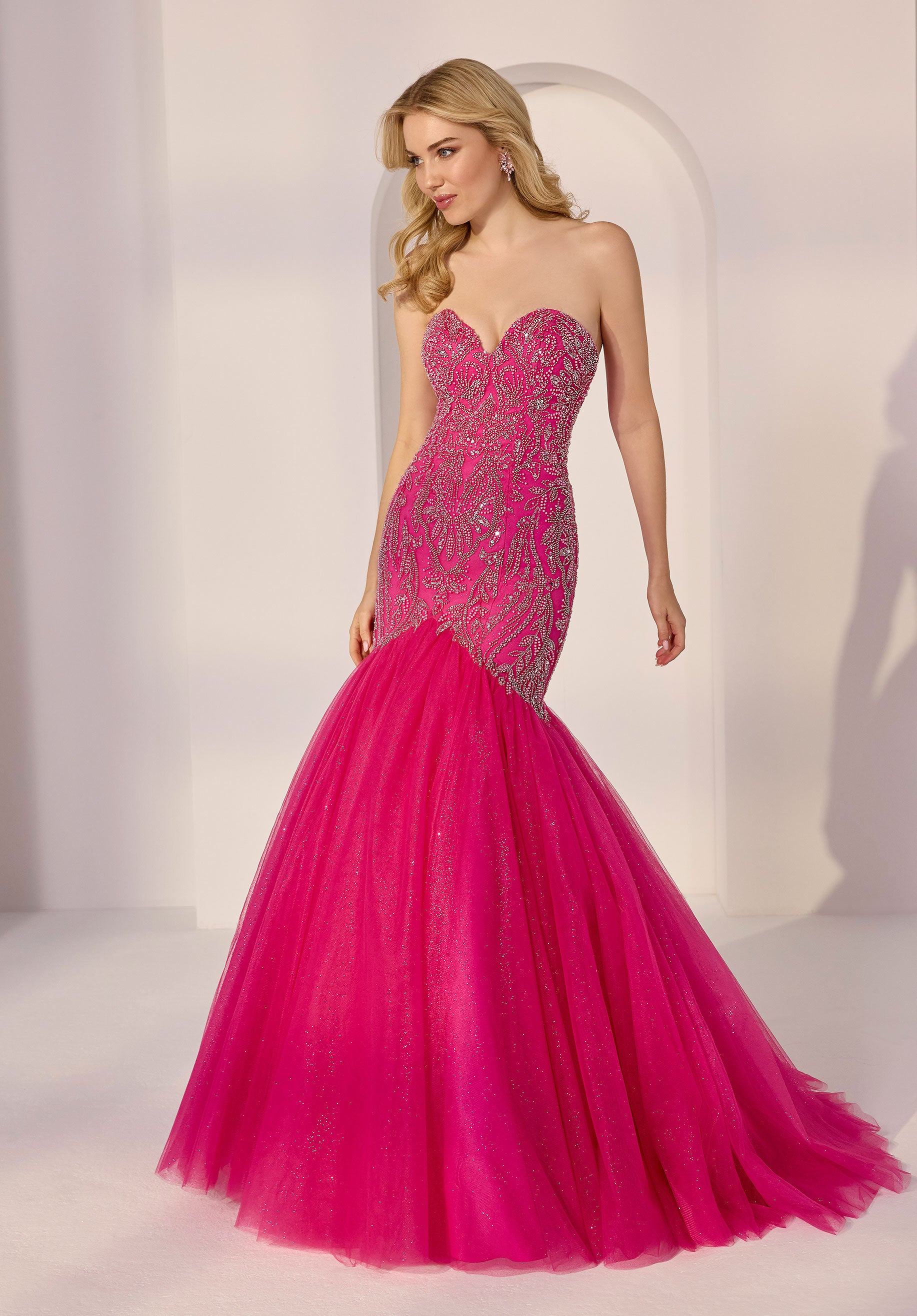 A woman stands in a white room wearing the Morilee 49203 Strapless Mermaid Prom Dress in hot pink, featuring a beaded bodice and a glitter tulle skirt.